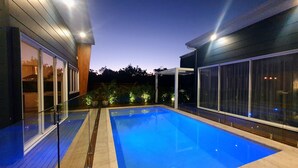 A modern outdoor swimming pool with glass railings and a wooden deck, surrounded by a dark-colored building with large windows. - The Family Beach House (Casuarina)
