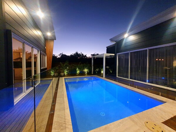 A modern outdoor swimming pool with glass railings and a wooden deck, surrounded by a dark-colored building with large windows.