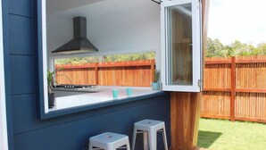 A modern outdoor kitchen with a built-in gas stove, a hood, and two bar stools. - The Family Beach House (Casuarina)