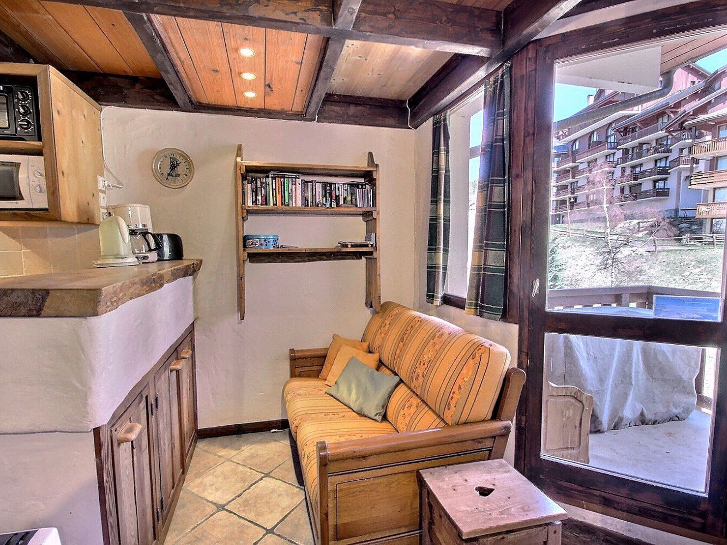 Una stanza accogliente con soffitto in legno, un angolo cottura con forno a microonde, una libreria, un'area salotto con cuscini e una finestra che si affaccia su edifici.