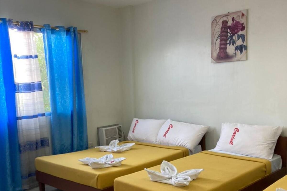 A hotel room with two single beds, each with a yellow bedspread and white pillows, and a blue curtain.