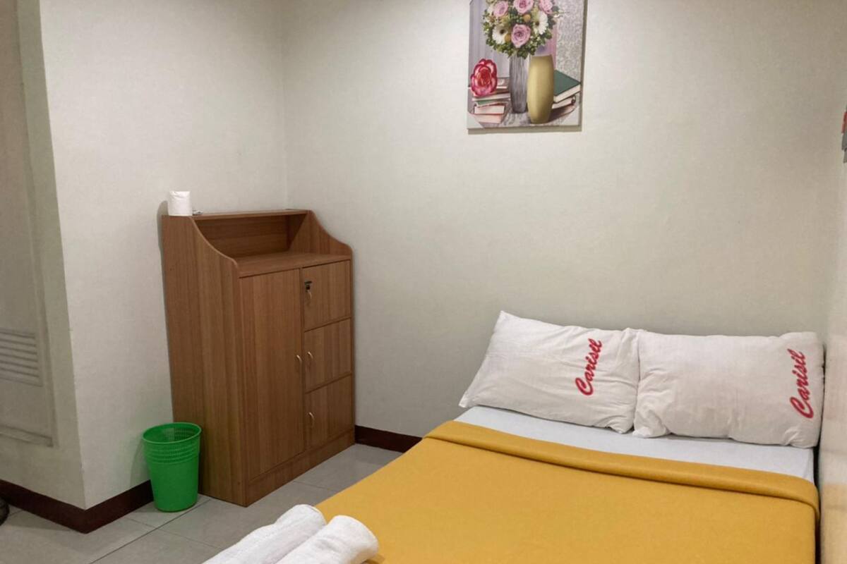A hotel room with a bed, a wooden wardrobe, a green trash bin, and a framed picture on the wall.