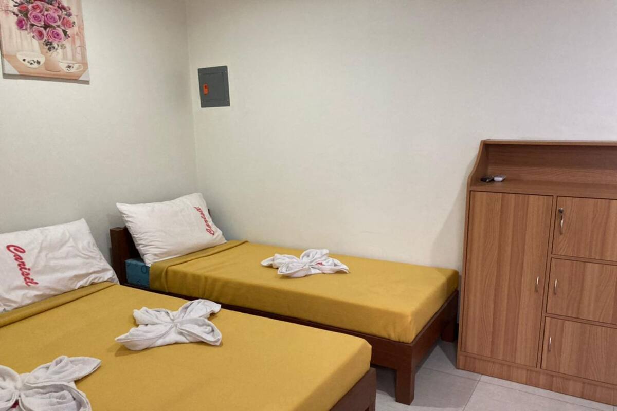 A hotel room with two single beds, each with a yellow bedspread and white pillows. There is a wooden wardrobe with two doors and a small table with a vase of flowers on the wall.