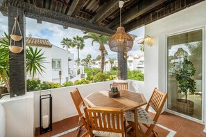 An outdoor patio with a wooden table and chairs, surrounded by plants and a view of neighboring buildings.