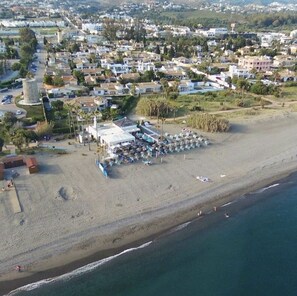 An aerial view of a coastal area with a beach, a row of houses, and a beachside restaurant with umbrellas.