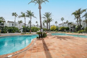 A swimming pool area with tiled flooring, palm trees, and a clear sky. - Costalita (Estepona)