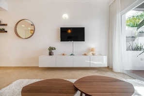 A modern living room with a flat-screen TV mounted on a white cabinet, a round mirror on the wall, and a two-seater coffee table.