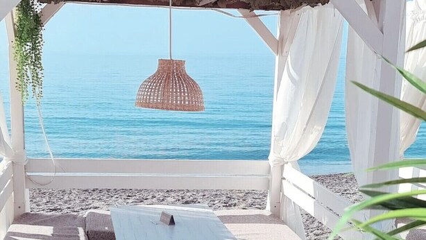 A beachside seating area with a view of the sea, a hanging light fixture, and a white canopy structure.