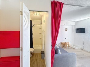 A modern bathroom with a shower, toilet, and a red curtain. There is a grey sofa and a small table in the living area with a TV mounted on the wall.