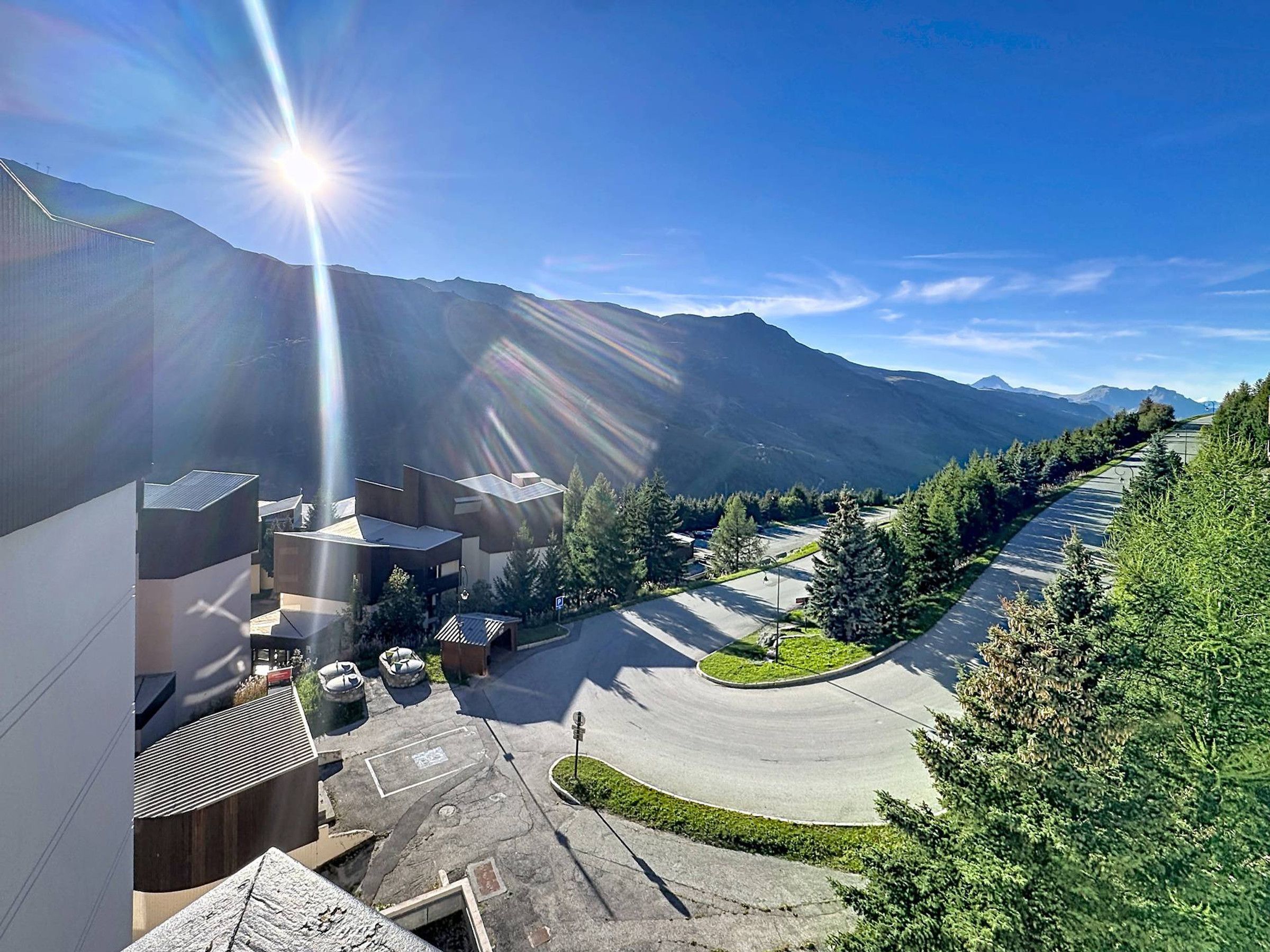 A parking area with a curved road, surrounded by greenery and mountains in the background.