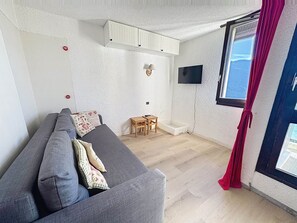 A room with a grey sofa, a small wooden table, a TV mounted on the wall, and a window with red curtains.