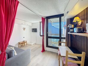 A room with a sofa, a small table, a TV, a window with curtains, and a kitchenette with a lamp and appliances.