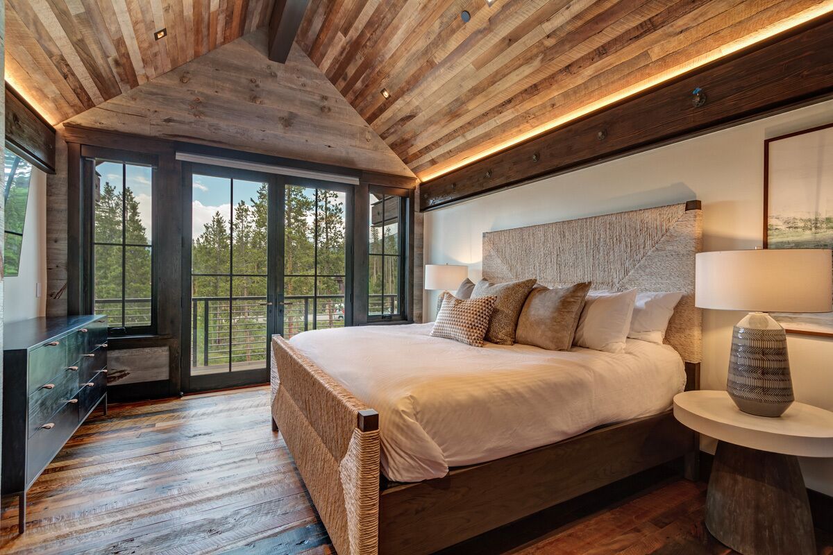 A bedroom with a large bed, wooden ceiling, and a view through large windows.
