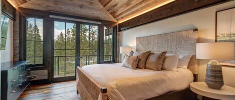A bedroom with a large bed, wooden ceiling, and a view through large windows.