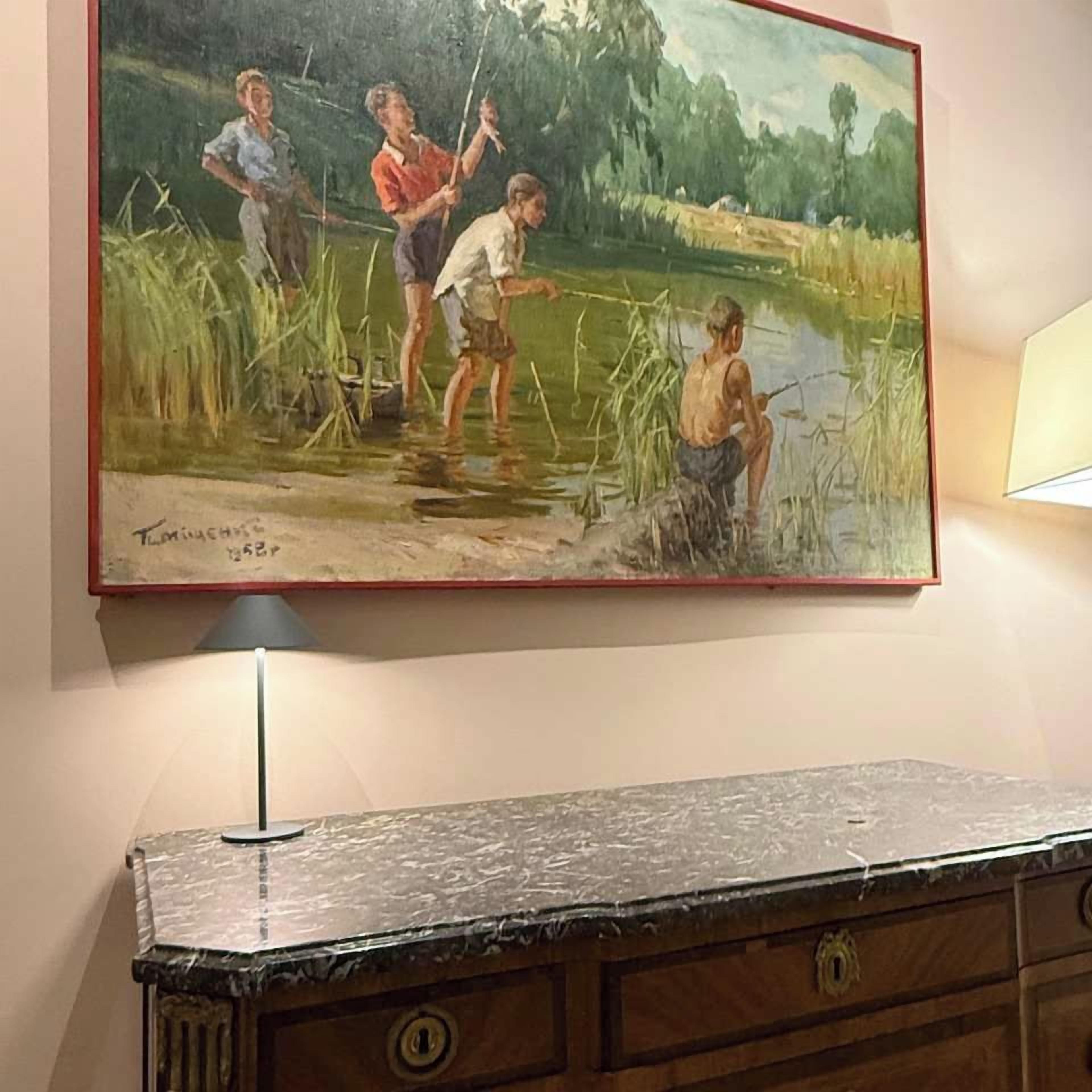 Un tableau représentant des personnes pêchant au bord d’un étang, avec une lampe et un meuble en bois en dessous.