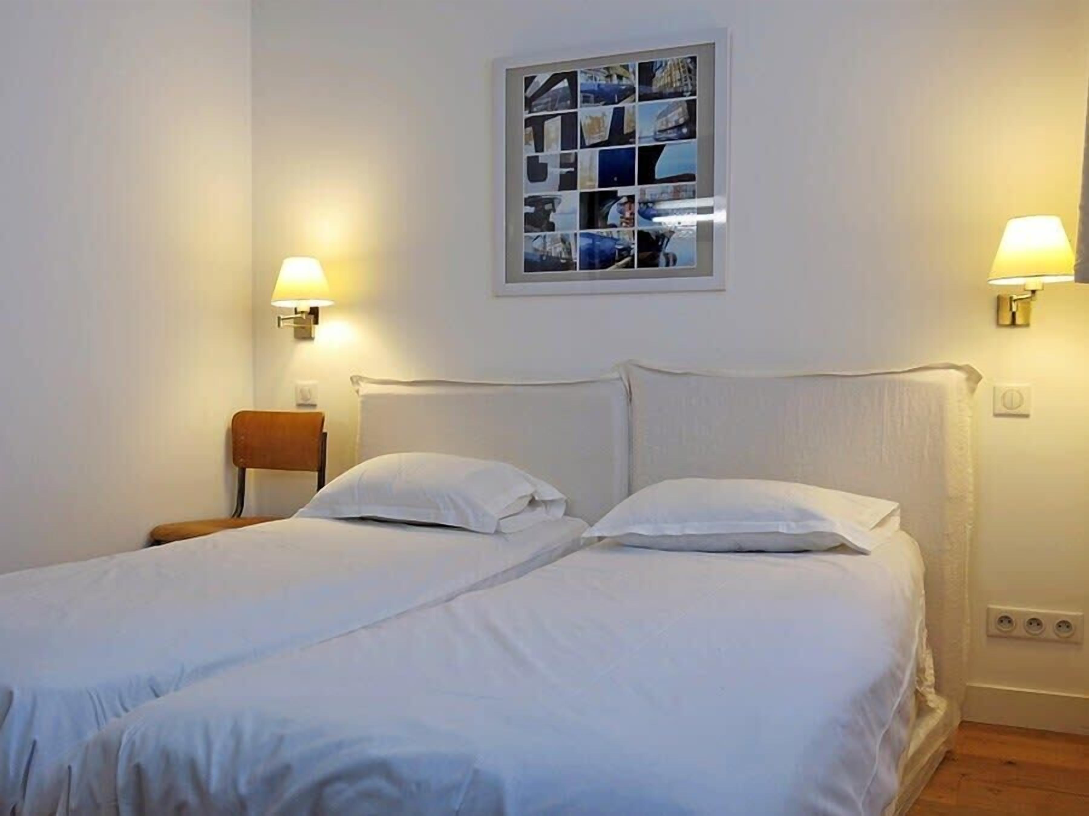 Un lit double avec du linge de lit blanc, deux lampes fixées au mur, un collage de photos encadrées accroché au mur et une chaise en bois.