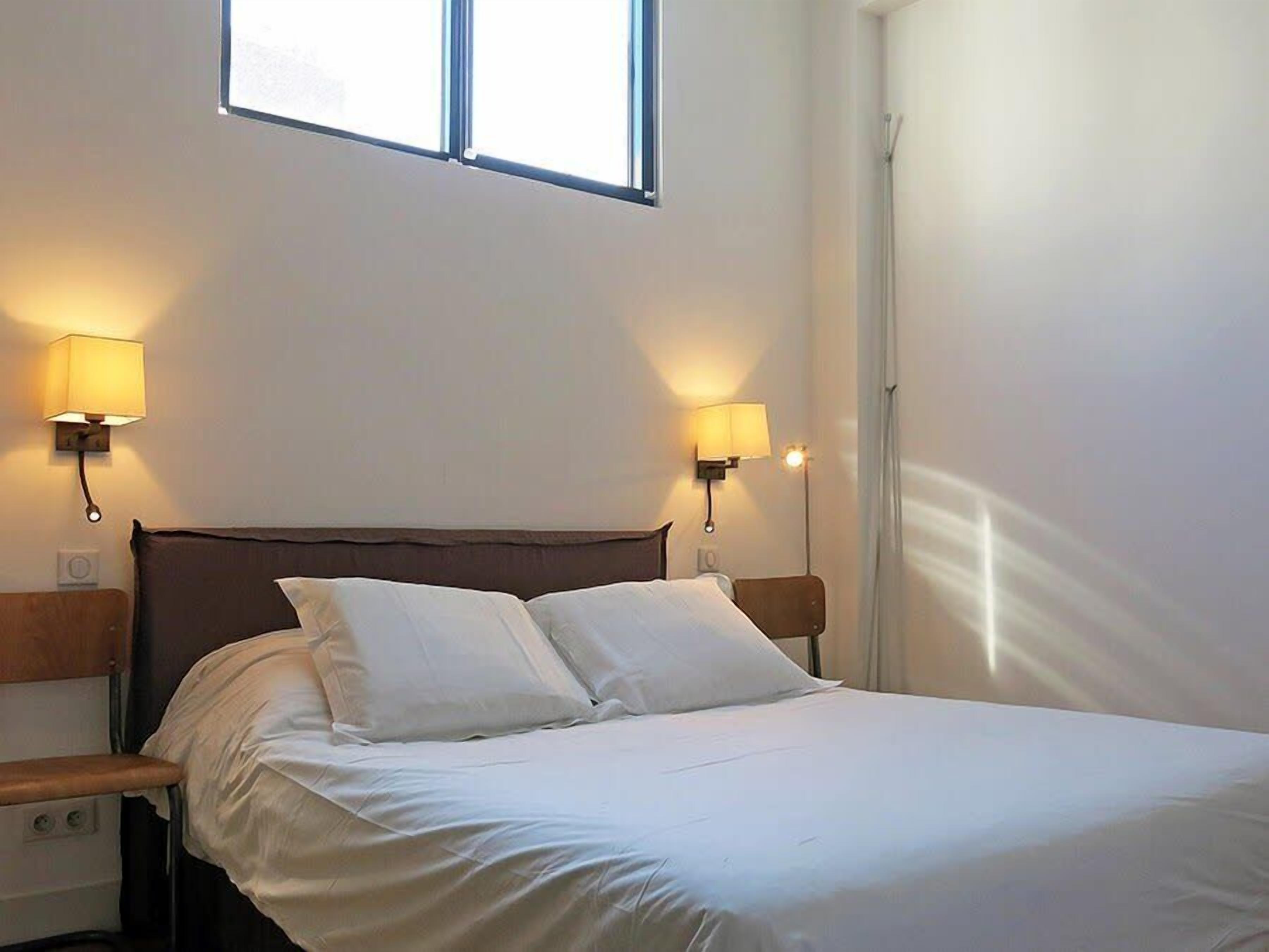 Uma cama bem arrumada com lençóis brancos, duas luminárias fixadas na parede e uma janela acima.