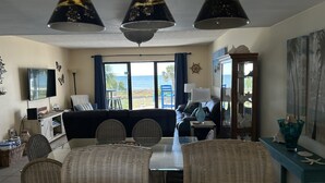 A living room with a TV, sofa, dining table, and chairs. There are three hanging lamps, a cabinet, and a view of the ocean. - SEASHELL ESCAPE - Pool & Beach Front 2 Bedroom sleeps 6 - Enjoy!!! (St. George Island)