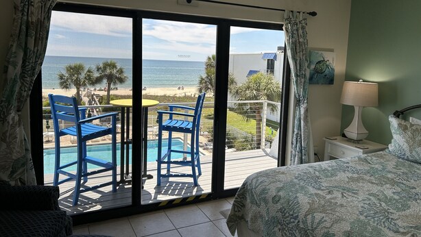 A room with a bed, a nightstand with a lamp, a view of the beach, and a pool area with chairs and a table. - SEASHELL ESCAPE - Pool & Beach Front 2 Bedroom sleeps 6 - Enjoy!!! (St. George Island)