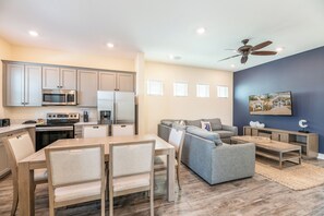 A modern kitchen with built-in appliances, a dining area with a wooden table and chairs, a gray sofa, and a ceiling fan.