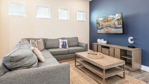 A living room with a grey sectional sofa, a coffee table, a TV mounted on a dark blue wall, and a wooden cabinet. - Sunrise Cottage near Disney with Margaritaville Resort & Island H20 Waterpark. (Kissimmee)