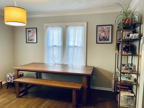 Un coin repas avec une table et un banc en bois, deux fenêtres ornées de rideaux légers et une bibliothèque contenant divers objets.