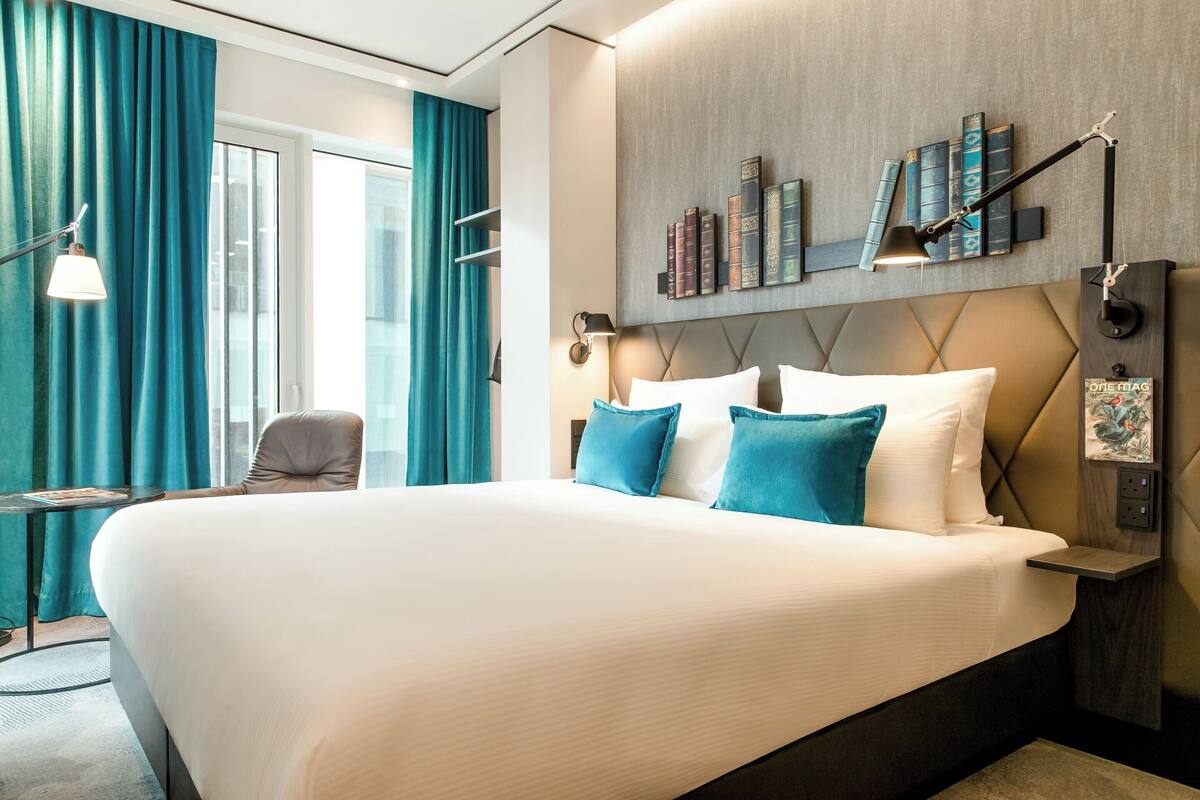 A large bed with white linens and blue pillows, a bedside table, a chair, a wall-mounted lamp, and a bookshelf with books.