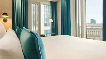Ein Hotelzimmer mit einem großen Bett, türkisblauen Vorhängen und Blick auf eine Stadtlandschaft durch die Fenster.