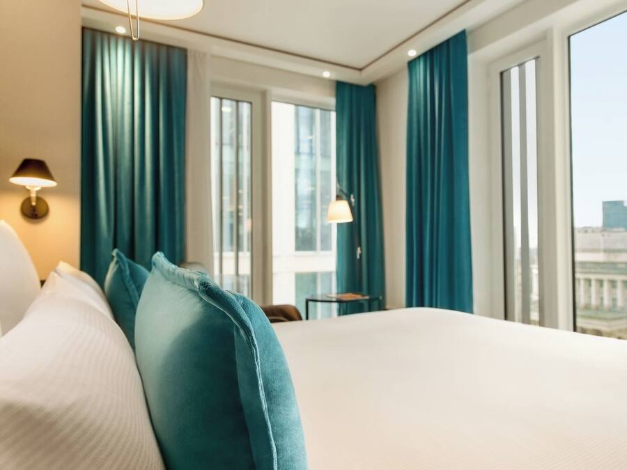 Habitación de hotel con una cama grande, cortinas color turquesa y vista a un paisaje urbano a través de las ventanas.
