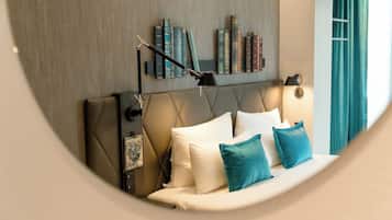 Una habitación de hotel moderna con un cabecero capitonado, una mesita de noche con lámpara, una estantería con libros y vistas al exterior a través de una ventana con cortinas.