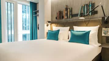 Ein Hotelzimmer mit einem großen Bett, türkis-farbenen Kissen, einem Bücherregal als Kopfteil und einem Nachttisch mit einer Lampe.