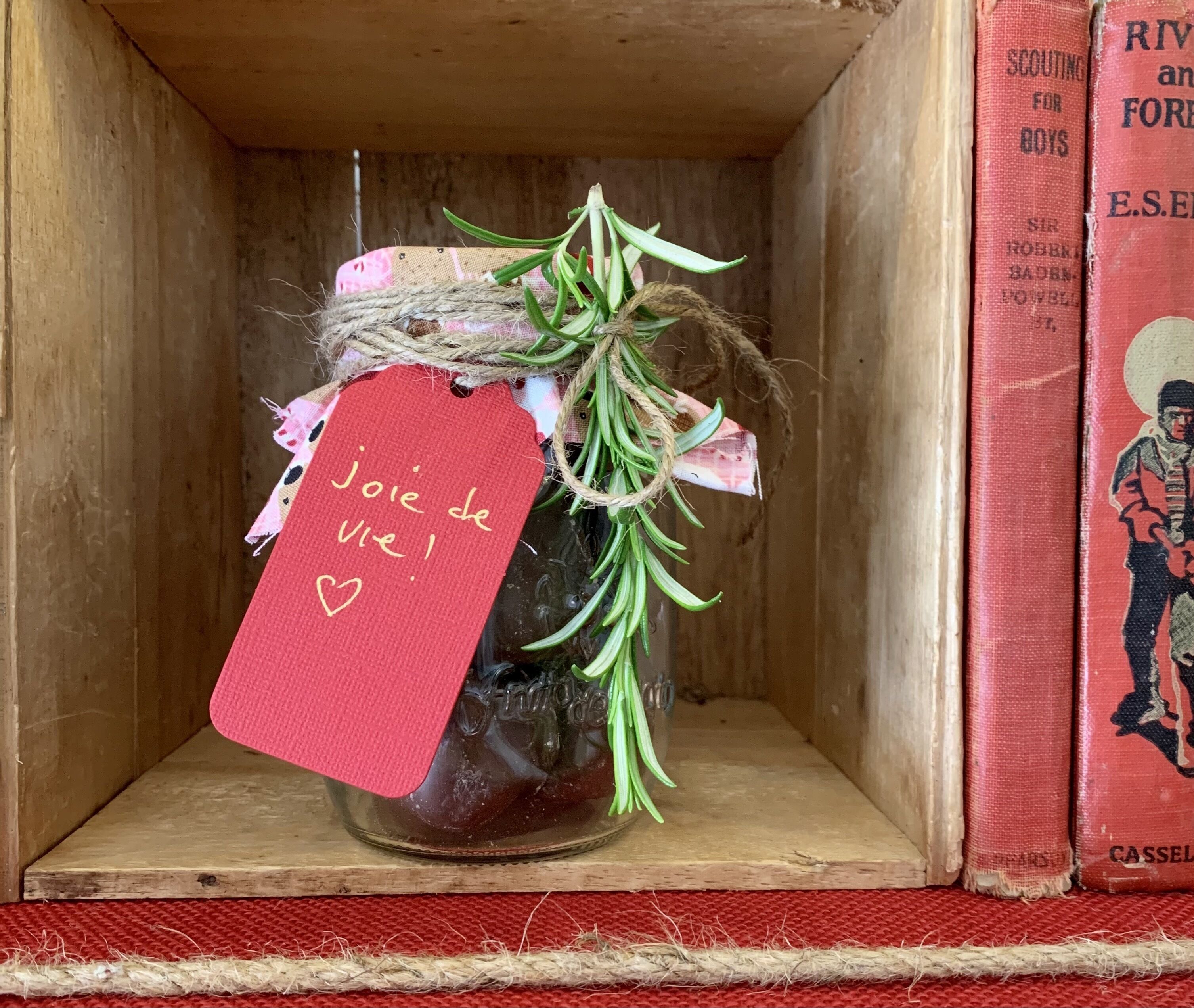 A small wooden box containing a glass jar with a tag reading "Joie de vivre!" and some green herbs.
