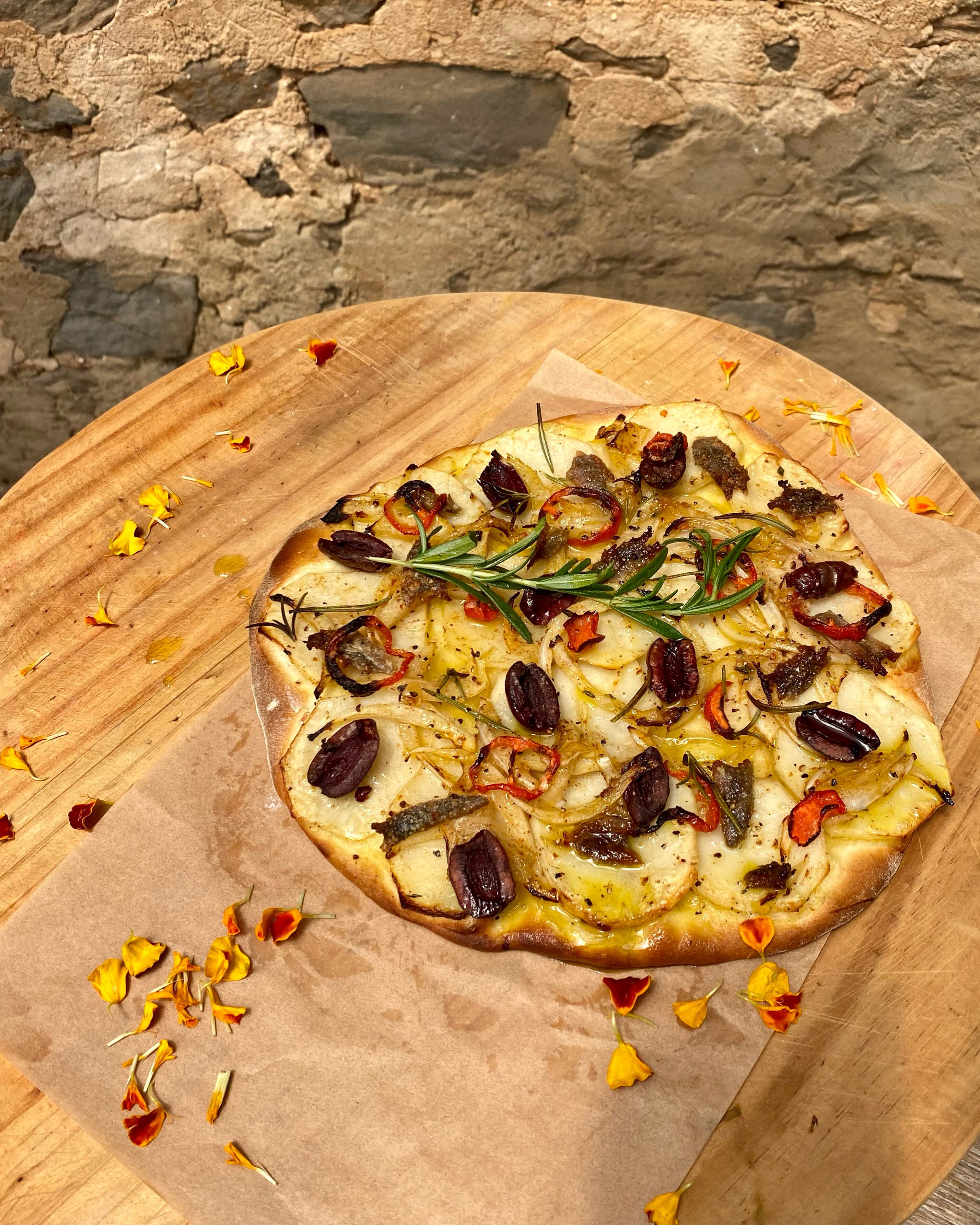 Eine Pizza auf einem Holzbrett, umgeben von verstreuten Blütenblättern.