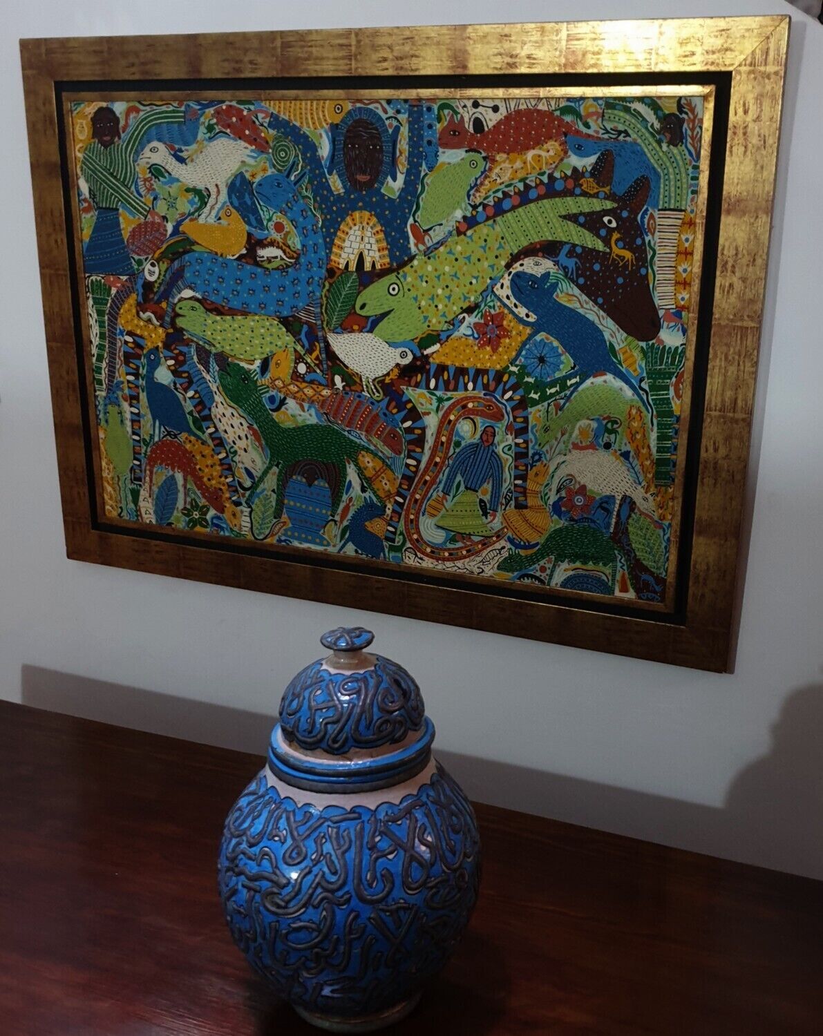 A framed colorful painting with a blue ceramic vase below it.
