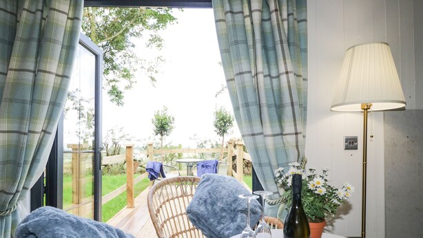 A cozy room with a wicker chair, a table with a bottle and glasses, and a view of the outdoors through large windows. - High Rigg Shepherd's Sunset (Brampton)