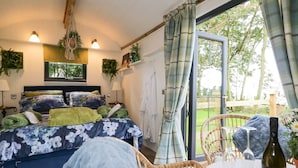 A bedroom with a bed, a bedside table, a chair, a table with a bottle, and a view of the outdoors. - High Rigg Shepherd's Sunset (Brampton)
