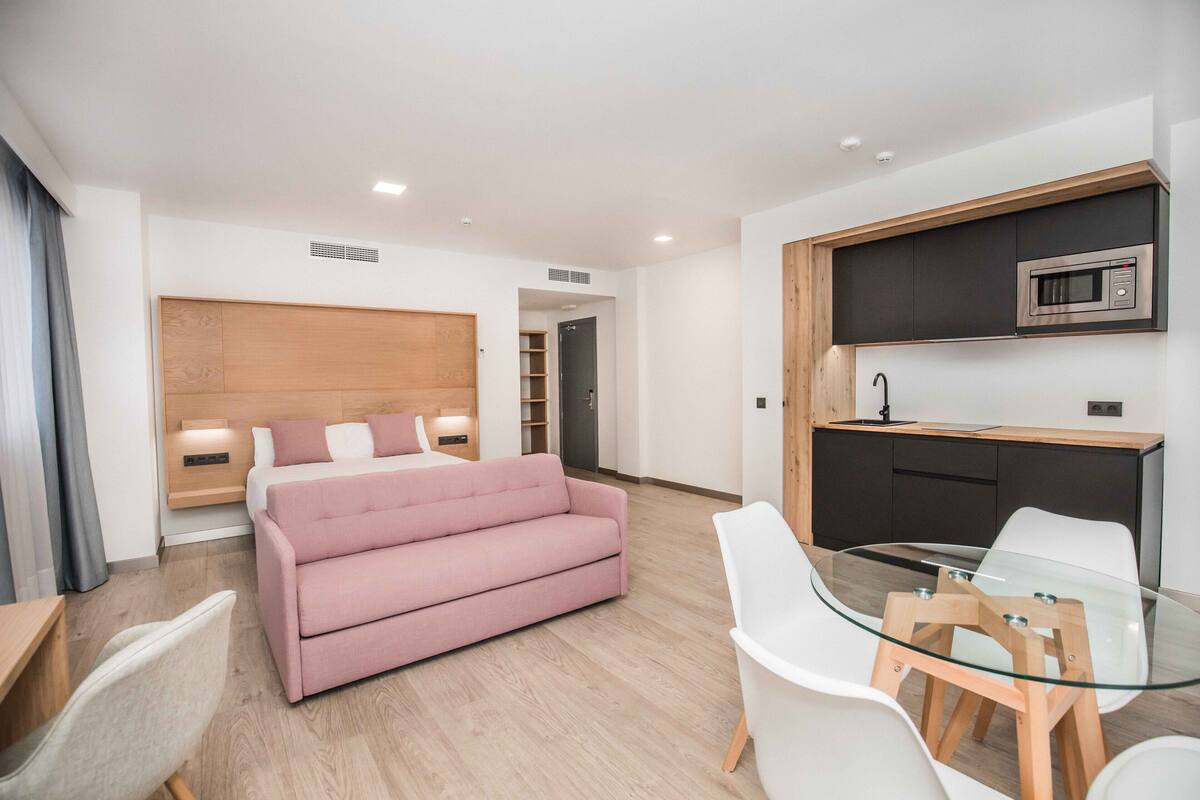 A modern hotel room with a bed, a pink sofa, a glass dining table, and a kitchenette.