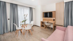 Een moderne woonkamer met een glazen eettafel, witte stoelen, een roze bank, een televisie en een houten bureau.