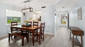 Un coin repas avec une table et des chaises en bois, un luminaire suspendu et une vue sur la cuisine et l’extérieur.