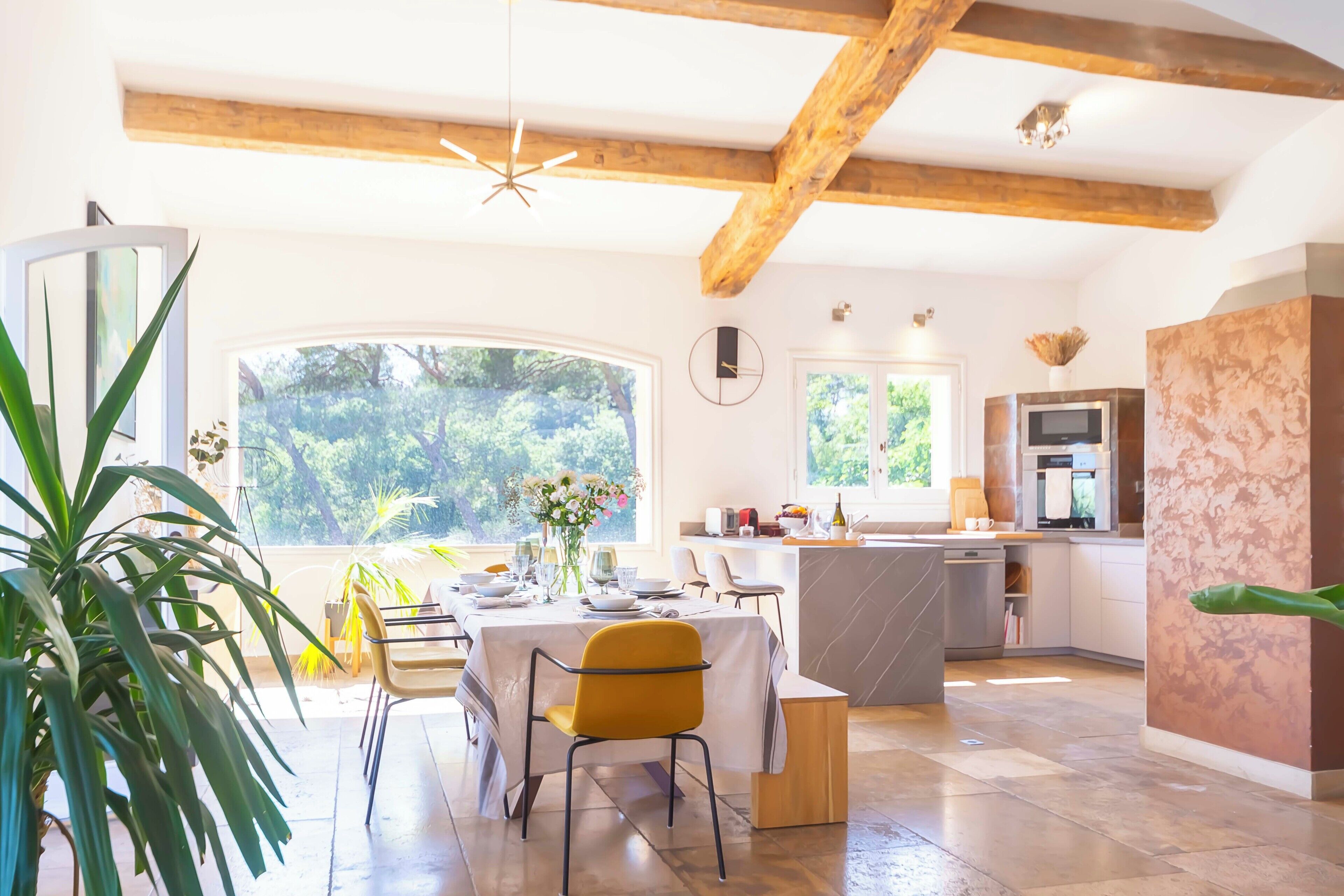 Une cuisine moderne dotée d’un grand îlot central, de poutres en bois et offrant une vue sur la verdure environnante.