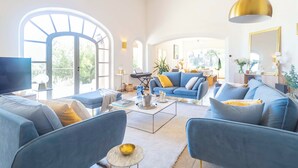 A spacious living room with a large arched window, a blue sofa set, a coffee table, and a television. - Panoramic 4-BR Villa near Aix w/Pool, AC - for 10 people (Le Tholonet (13100))