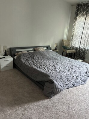 Un lit bien fait, avec une couette grise, une table de chevet avec une lampe et une chaise agrémentée d’un coussin à motifs.