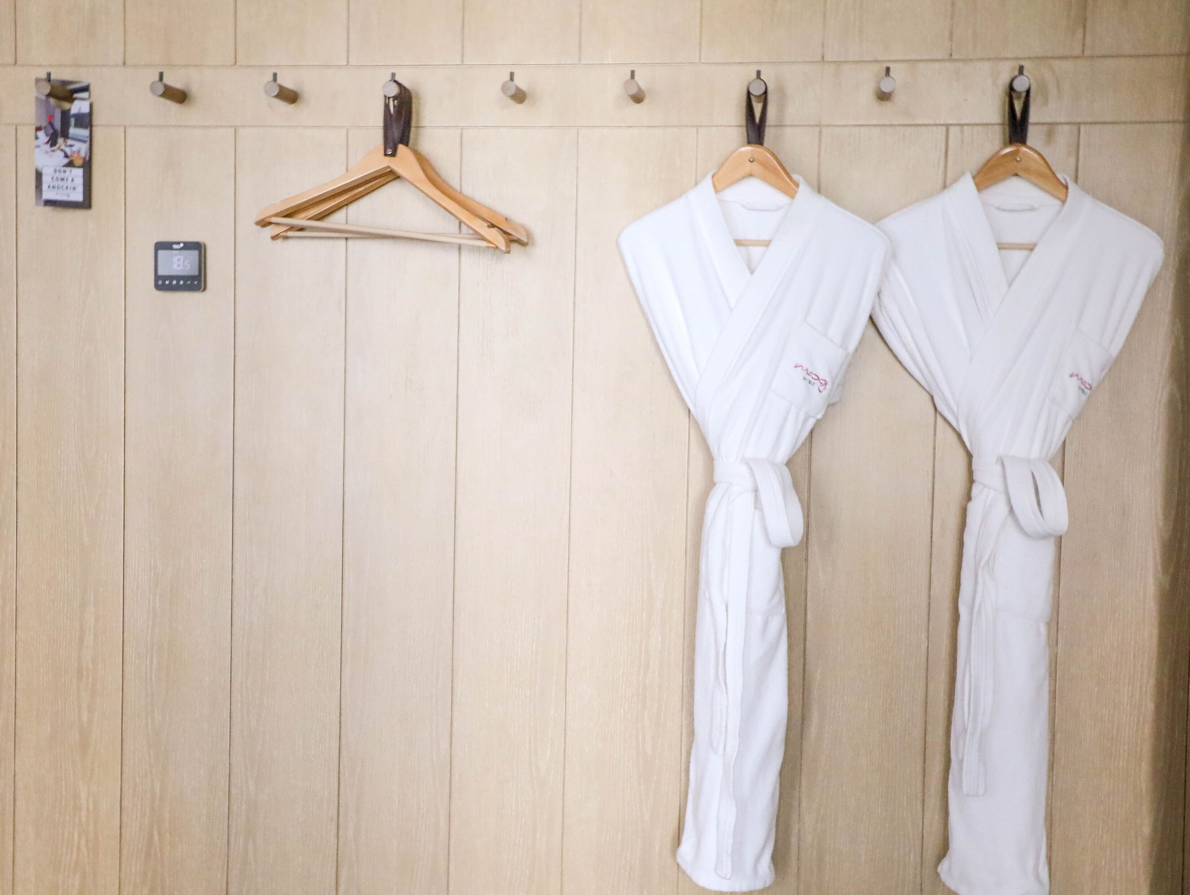 A wooden wall with two white robes hanging on wooden hangers, a small digital thermometer, and a framed picture.