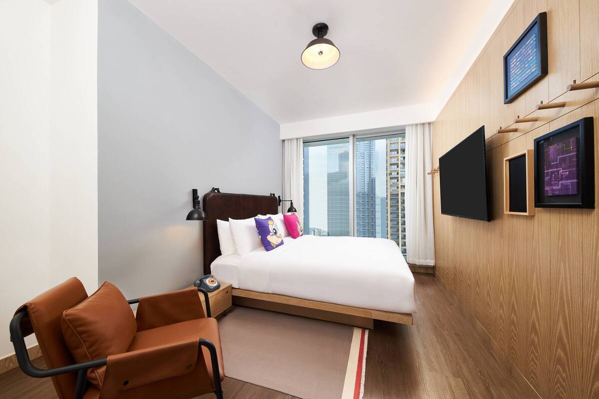 Moderni hotellihuone, jossa on sänky, työpöytä, tuoli, suuri ikkuna, josta on näkymä kaupunkiin, sekä seinälle asennettu taulutelevisio.