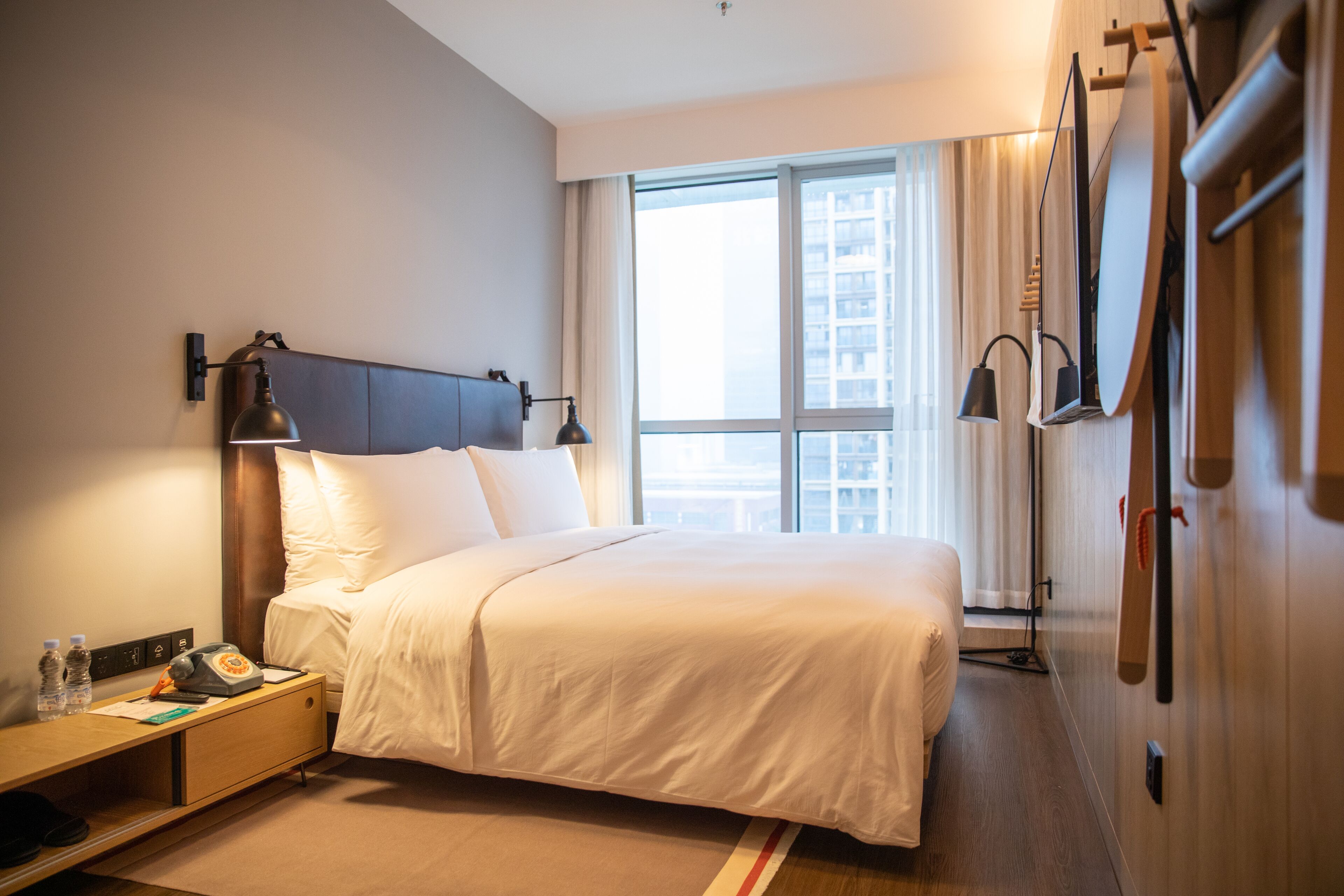 A hotel room with a large bed, bedside lamps, a nightstand, a window with curtains, and a view of the city.