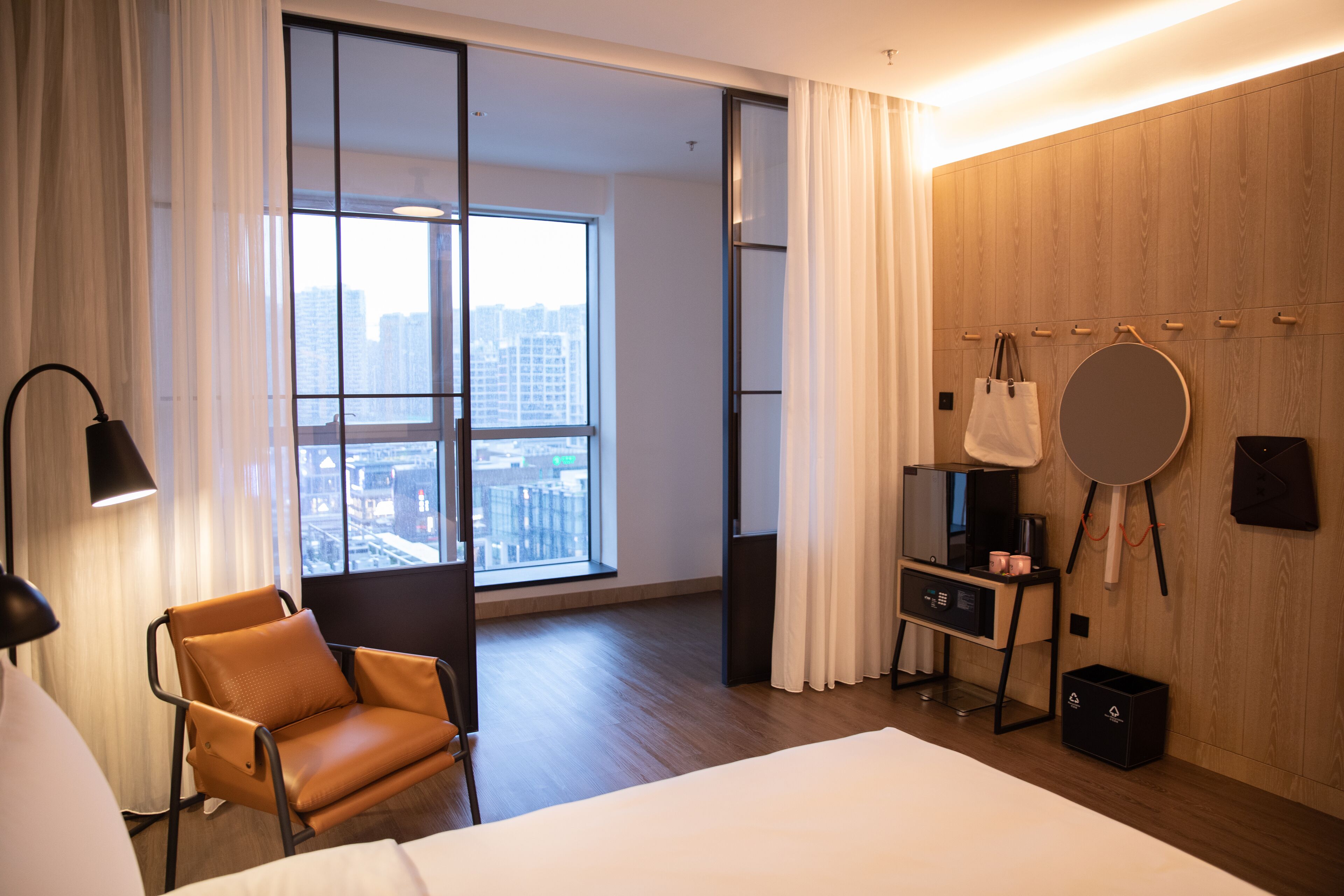 A modern hotel room with a large window, a bed, a chair, a vanity with a mirror, and a wall-mounted coat rack.