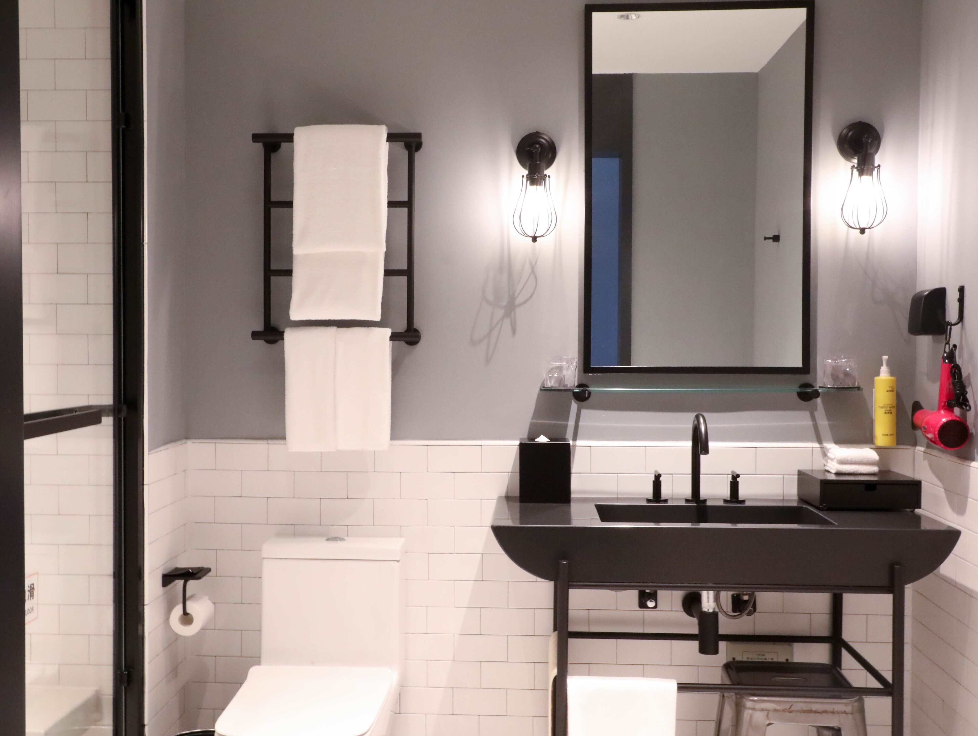Une salle de bain moderne avec des toilettes, un lavabo et un miroir. On y trouve des serviettes suspendues à un porte-serviettes, un distributeur de savon et un sèche-cheveux fixé au mur.