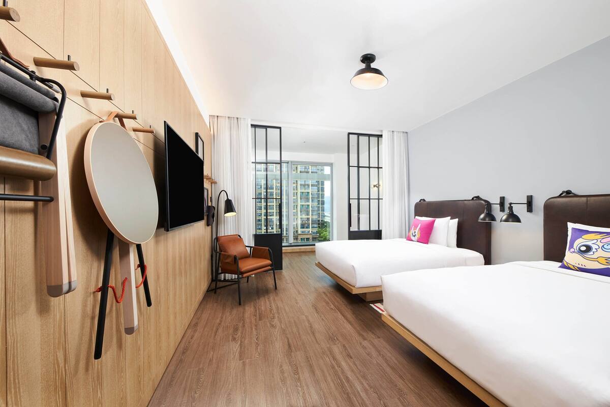 Hotellihuone, jossa on kaksi sänkyä, televisio, tuoli ja suuri ikkuna, josta avautuu näkymä kaupunkiin.