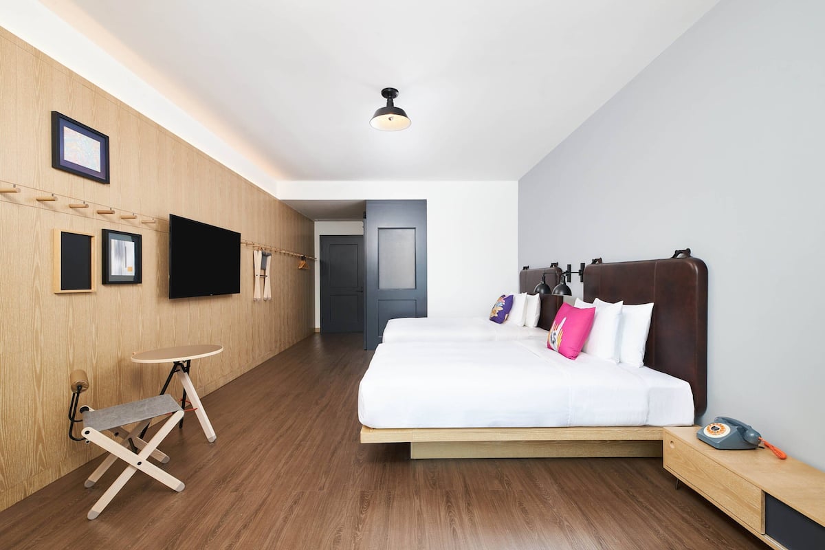 Moderni hotellihuone, jossa on puulattia, valkoisilla vuodevaatteilla varustettu sänky, yöpöytä, jossa on puhelin, ja pieni työpöytä tuolilla.