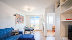 A living room with a blue sofa, a dining table with chairs, a TV on a stand, and a view of the outside through a glass door. - PASSY AZUR AP4242 - Vue mer à 10 min de la plage (Niza)
